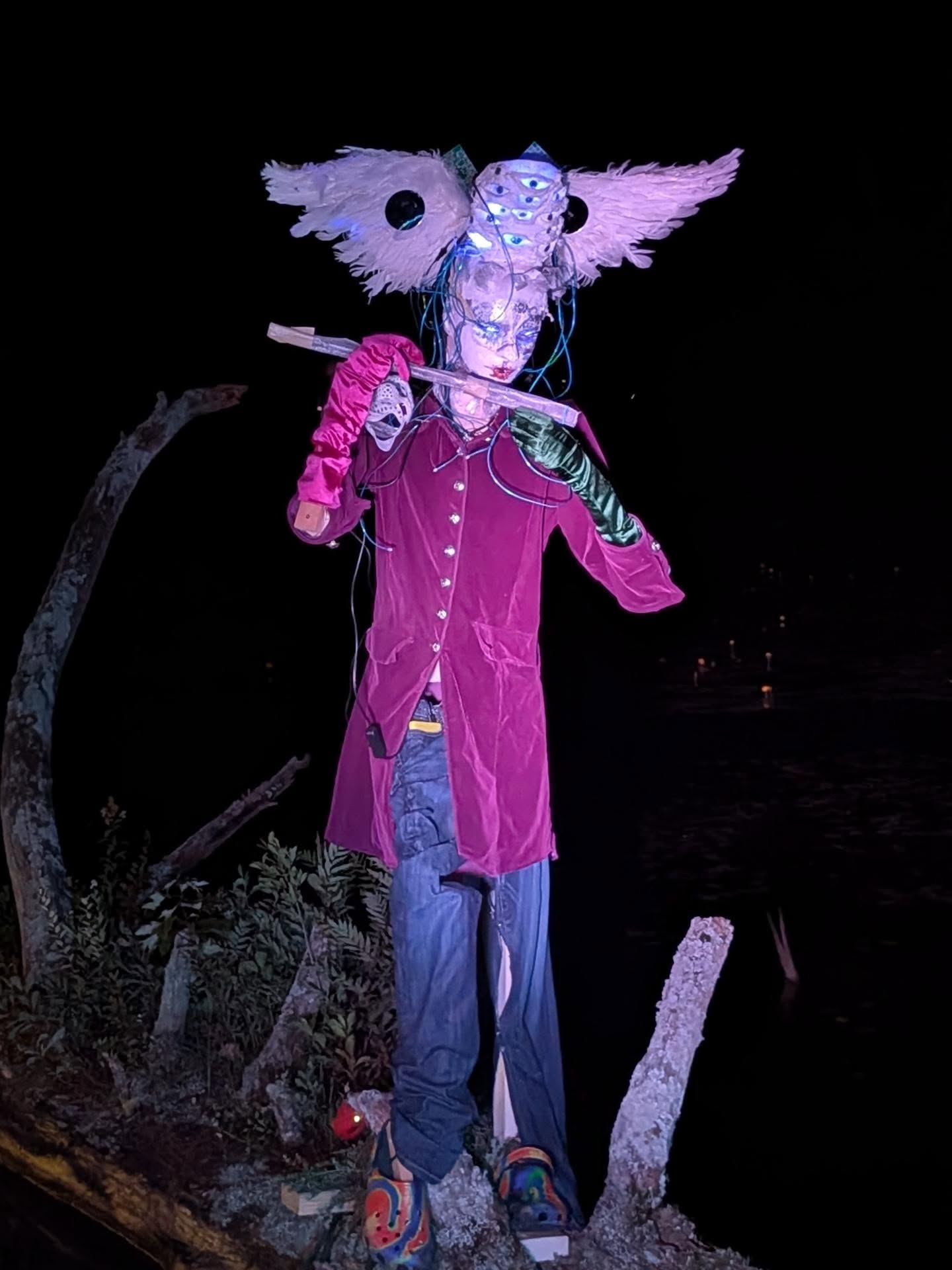 The PiperGPT sculpture: a many-eyed, winged half-machine figure in a magenta velvet coat playing a wooden flute at night