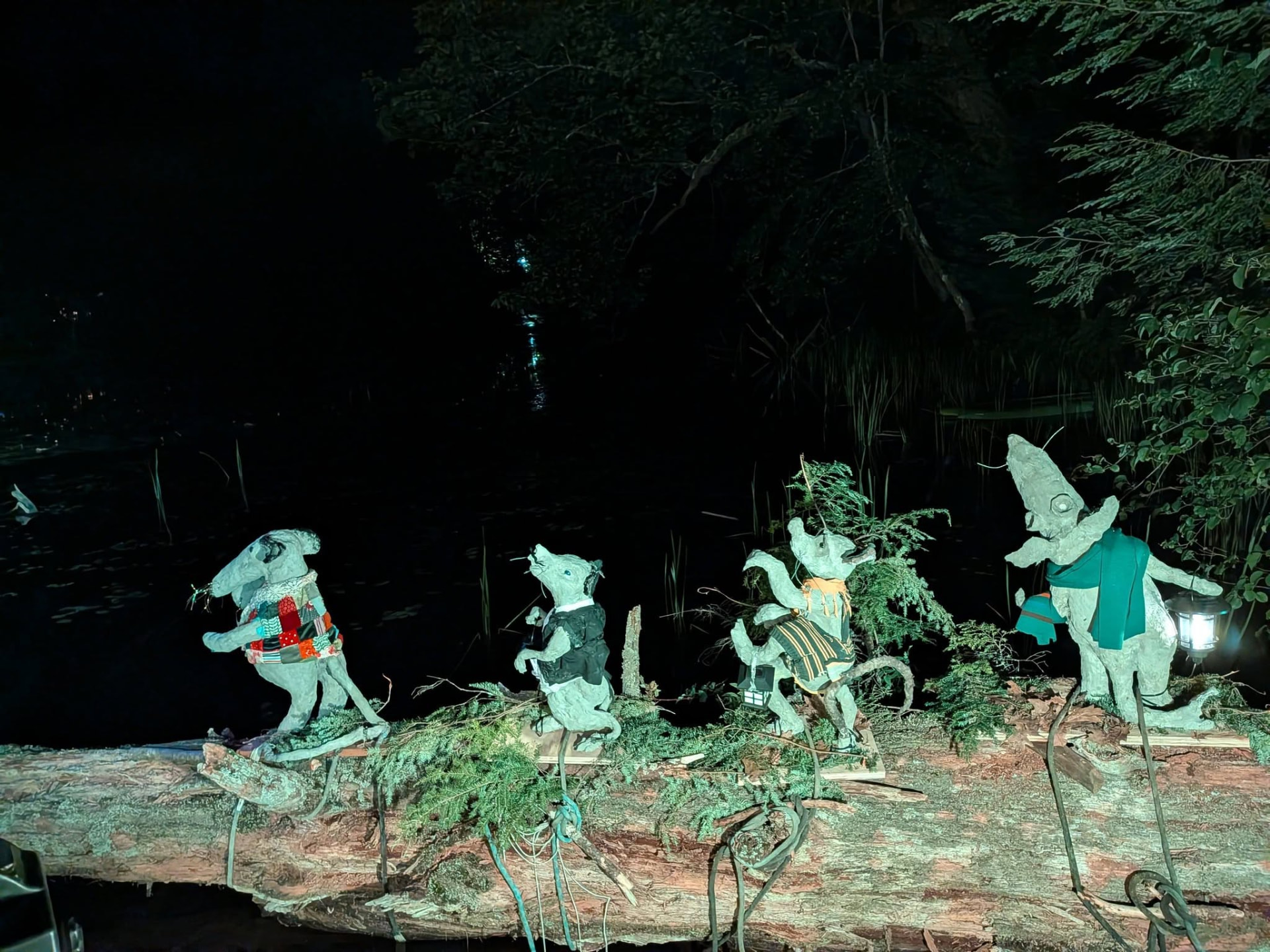 Four hand-sculpted rats in coats and lanterns walking along a fallen log above dark water, lit in teal light